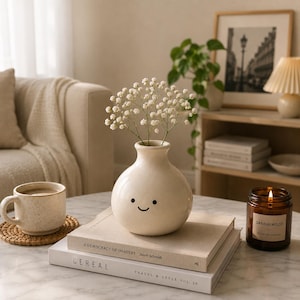 May include: A cream-colored vase with a smiling face holds white flowers, resting on books labeled "CEREAL" and "TRAVEL & STYLE VOL. 20." A mug, a candle labeled "SANDALWOOD," and a framed picture complete the cozy, neutral-toned scene.