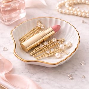May include: A gold lipstick tube and several gold hairpins with pearl and crystal accents rest in a white, shell-shaped dish with a gold rim. A string of pearls and a perfume bottle are in the background.