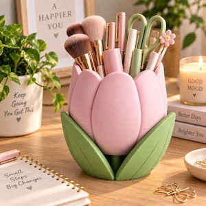 May include: A pink tulip-shaped desk organiser with green leaves at the base, holding various makeup brushes, pens, and light green scissors. The organiser is made of smooth material and features a flower design with petals and leaves.