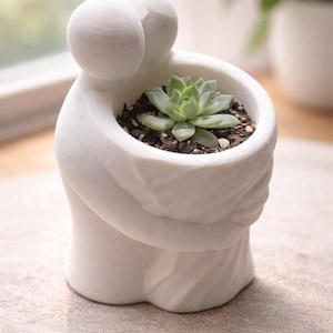 May include: A white ceramic planter shaped like two figures embracing, holding a small succulent plant. The planter has a smooth finish and is filled with soil. The succulent has green leaves.