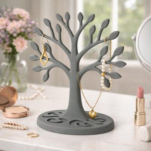 Op de afbeelding: Een grijze boomvormige sieradenorganizer met bladvormige takken. Gouden oorbellen, een parelketting en een ketting met een hartvormige hanger hangen aan de takken. Een gouden ring en een compactpoeder liggen op de tafel.