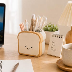 Può includere: Un portapenne in ceramica a forma di fetta di pane con un volto sorridente. Il portapenne è pieno di penne e matite. Un piccolo blocco note con le parole "Focus. Plan. Do. Repeat" è accanto al portapenne.