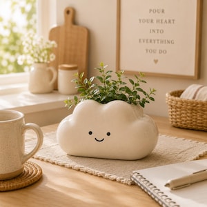 May include: A white ceramic cloud-shaped planter with a smiling face, holding green plants. The planter sits on a beige placemat on a wooden table. A mug, a coaster, and a notebook are also visible.