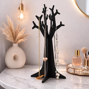 May include: A black tree-shaped jewelry organizer displaying necklaces with gold and pearl accents. The organizer is on a marble surface, with a vase of pampas grass and a vanity mirror in the background.