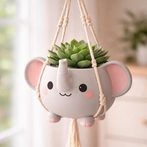 Puede incluir: Un macetero colgante gris con forma de elefante y una planta suculenta verde. El macetero tiene detalles rosas y una cara sonriente. Cuelga de una cuerda de macramé.