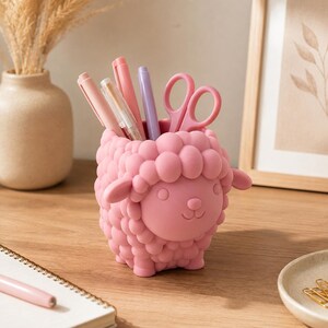 May include: A pink sheep-shaped desk organizer featuring a textured, rounded wool design. The container holds several pens in shades of pink and purple, along with a pair of matching pink scissors, providing a functional storage solution for stationery.