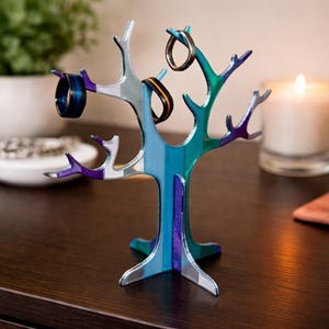 May include: A colorful tree-shaped ring holder with a striped design in shades of blue, green, and purple. Several rings are displayed on the branches. A lit candle and a plant are in the background.