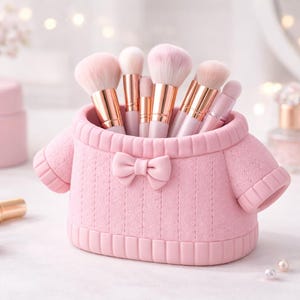 May include: A pink makeup brush holder shaped like a sweater, with a bow detail. The holder contains several makeup brushes with pink bristles and gold-colored handles. The background includes other beauty products.