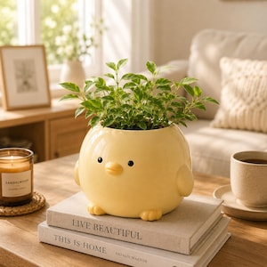 May include: A yellow ceramic chick-shaped planter with a green plant inside. The planter has black eyes, a yellow beak, and small feet. It sits atop two books with the titles "LIVE BEAUTIFUL" and "THIS IS HOME".