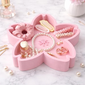 May include: A pink, flower-shaped jewelry tray filled with accessories. The tray holds a pink scrunchie, hair clips, a pearl necklace, earrings, and a small perfume bottle. Additional pearls and hair pins are scattered around the tray.