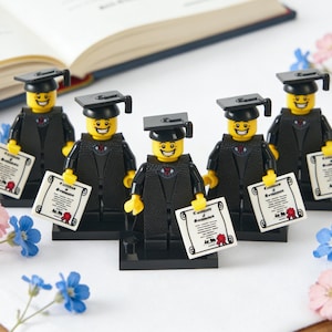 May include: Five Lego figures dressed in graduation attire, holding diplomas. Each figure wears a black graduation gown and mortarboard. The figures are yellow with smiling faces. Pink and blue flowers and an open book are in the background.