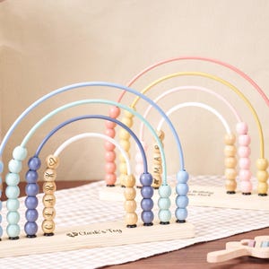 May include: Two wooden rainbow-shaped abacus toys with pastel-colored arches and beads. One is personalized with the name "Clark" and the other with "Mia". The base of each toy is engraved with "Clark's Toy".