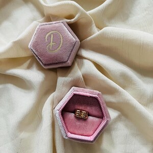 Könnte beinhalten: Zwei sechseckige, rosa Samt-Ringboxen. Eine Box ist mit einem goldenen Buchstaben "D" auf der Oberseite geschlossen. Die andere Box ist offen und zeigt einen goldenen Ring. Die Boxen liegen auf einem cremefarbenen, strukturierten Stoff.