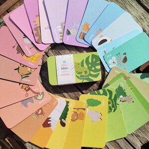 Könnte beinhalten: Ein Set aus 25 rechteckigen Karten mit bunten Tierillustrationen und deutschem Text, fächerförmig angeordnet um eine zentrale Schachtel mit der Aufschrift 25 Mini-Bibelkarten, einem grünen Chamäleon und einem Monstera-Blatt-Motiv.