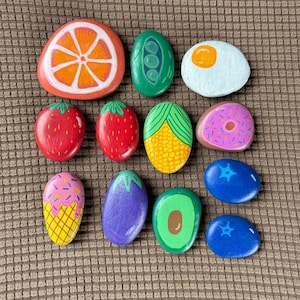 Pode incluir: Uma coleção de pedras pintadas retratando vários alimentos. As pedras incluem uma fatia de laranja, ervilhas, um ovo frito, morangos, milho, um donut, um sorvete, uma berinjela, um abacate e mirtilos. As cores são brilhantes.