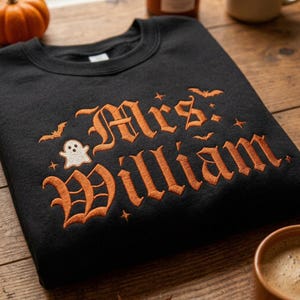 May include: A black crewneck sweatshirt featuring orange embroidered text that reads Mrs. William. The design includes a small white ghost, several orange bats, and tiny star accents surrounding the lettering in a gothic-style font.