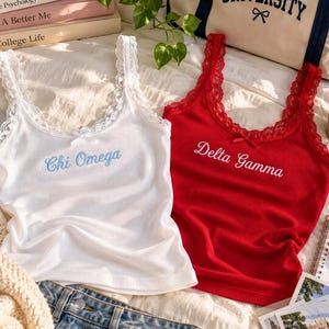 Könnte beinhalten: Zwei gerippte Tanktops mit Spitzenbesatz und kleinen Schleifen am Ausschnitt. Das weiße Tanktop zeigt den blauen Sticktext Chi Omega, während das rote Tanktop den weißen Sticktext Delta Gamma zeigt. Beide Kleidungsstücke haben einen Rundhalsausschnitt.