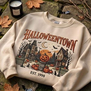 May include: A beige crewneck sweatshirt featuring a graphic print with the text HALLOWEENTOWN EST. 1998. The illustration depicts two houses, a crescent moon, a large orange pumpkin on a pedestal, smaller pumpkins, bats, and black cats.