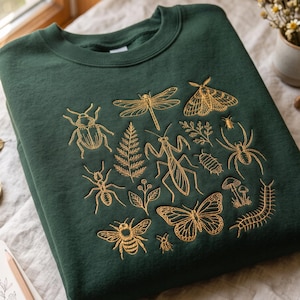 May include: A dark forest green crewneck sweatshirt featuring gold-colored embroidered illustrations of various insects, including a dragonfly, moth, mantis, spider, beetle, bee, and centipede, alongside botanical elements like a fern and mushrooms.