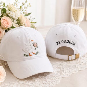 May include: Two white baseball caps. One cap features an embroidered goose wearing a veil and holding a bouquet of flowers. The other cap displays the embroidered date 21.03.2026 above the adjustable strap with a gold-toned metal buckle.