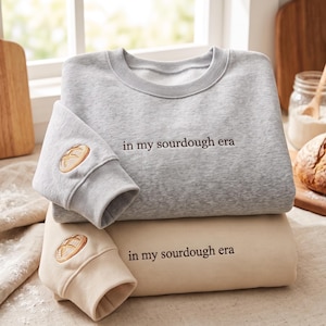 May include: A light gray crewneck sweatshirt and a beige crewneck sweatshirt, each featuring the embroidered text "in my sourdough era" on the chest and a small embroidered sourdough bread loaf design on the sleeve cuff with ribbed trim.