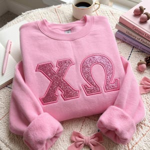 Puede incluir: Una sudadera de cuello redondo en color rosa claro con las letras griegas Chi y Omega en un acabado de purpurina rosa oscuro con un borde cosido. La sudadera tiene mangas largas con puños acanalados y un interior de forro polar suave.