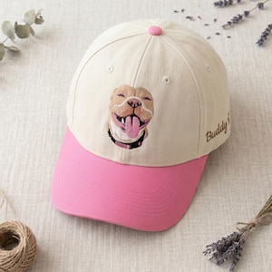 Pode incluir: Um boné de beisebol com copa cor de creme e aba rosa. A frente apresenta um cão bege bordado com a língua de fora. A palavra Buddy está bordada em castanho na lateral do boné. O boné tem um pequeno botão rosa no topo.