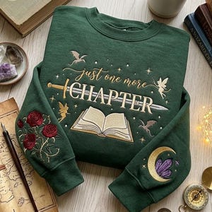 以下が含まれることがあります： 「just one more CHAPTER」という金色の刺繍文字が、開いた本、剣、小さなドラゴン、星の上に施されたダークグリーンのクルーネックトレーナー。袖には赤いバラと、クリスタルがあしらわれた金色の三日月の刺繍が施されています。