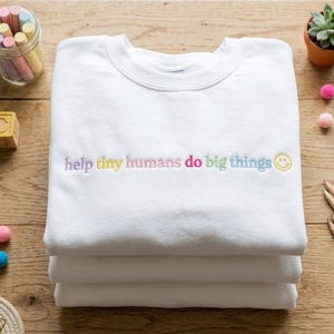 Puede incluir: Una sudadera blanca de cuello redondo con el texto bordado "help tiny humans do big things" en letras pastel multicolores, seguido de un pequeño icono de cara sonriente amarilla centrado en el pecho de la prenda.