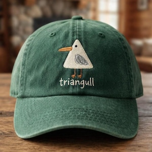 Puede incluir: Una gorra de béisbol verde bosque con un bordado que muestra una gaviota triangular blanca con pico y patas naranjas. Debajo del ave, la palabra triangull está bordada en hilo blanco. La gorra tiene una visera curva y costuras.