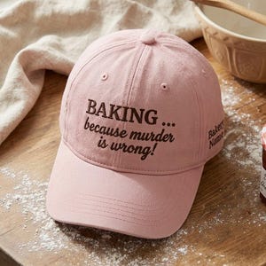 Op de afbeelding: Een lichtroze honkbalpet met donkerbruine geborduurde tekst die luidt BAKING... because murder is wrong! De zijkant van de pet bevat extra geborduurde tekst die Bakery Name weergeeft in een donkerbruin lettertype.