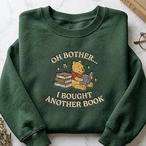 Puede incluir: Una sudadera de cuello redondo en color verde oscuro con un bordado de un oso amarillo sentado junto a una pila de libros mientras lee. El texto dice "OH BOTHER... I BOUGHT ANOTHER BOOK", con pequeños detalles de estrellas y una abeja.