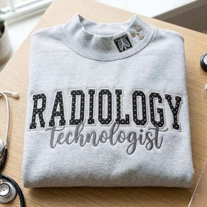 以下が含まれることがあります： ライトグレーのクルーネックトレーナー。黒地に白の水玉模様の「RADIOLOGY」という刺繍と、グレーの筆記体で「technologist」という刺繍が施されています。襟元には放射線マークと肋骨のグラフィックアイコンが刺繍されています。