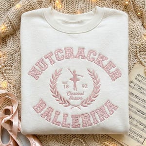 May include: A cream-colored crewneck sweatshirt featuring pink embroidered text that reads NUTCRACKER BALLERINA. The design includes a central ballerina silhouette, a laurel wreath, and the text EST 1892 Classical Season in pink thread.