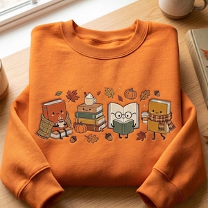 Puede incluir: Una sudadera naranja de cuello redondo con una ilustración gráfica de libros antropomórficos. El diseño incluye libros con caras, una manta de cuadros, una bufanda, una taza de café, una calabaza y hojas de otoño dispersas por el centro.
