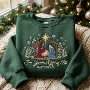 Pode incluir: Uma camisola de gola redonda verde-escura com uma cena de natividade bordada, incluindo uma estrela, pinheiros e flocos de neve. O texto The Greatest Gift of All e MATTHEW 1:21 está bordado a branco no centro do peito.