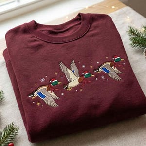 May include: A maroon crewneck sweatshirt featuring an embroidered design of three mallard ducks in flight. Each duck wears a small red Santa hat and a red scarf. The embroidery is accented with small white snowflakes and gold stars.