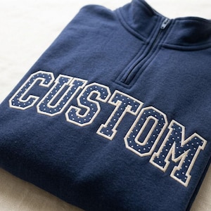 Peut inclure: Un pull bleu marine à fermeture éclair quart de longueur avec le mot CUSTOM en grandes lettres de style bloc sur la poitrine. Les lettres sont soulignées d'une couleur beige clair et remplies d'un tissu bleu marine à pois blancs.