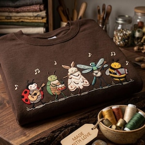 Puede incluir: Una sudadera marrón oscuro con un bordado de cinco insectos tocando instrumentos musicales, incluyendo una mariquita con guitarra, un escarabajo con tambor, una polilla con flauta, una libélula con banjo y una abeja con un acordeón.