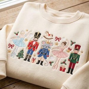 May include: A cream-colored crewneck sweatshirt featuring colorful embroidery of two nutcrackers, two ballerinas, a mouse wearing a crown, a rocking horse, Christmas trees, gifts, holly, and bows arranged across the chest area of the garment.