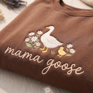 May include: A brown crewneck sweatshirt featuring an embroidered design of a white goose and a yellow gosling surrounded by white daisies with green stems. The text mama goose is embroidered in a cursive font below the goose and gosling.