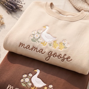 May include: A beige crewneck sweatshirt featuring an embroidered design of a white goose, two yellow goslings, and white daisies with green stems. The text "mama goose" is embroidered in brown script below the goose and goslings illustration.