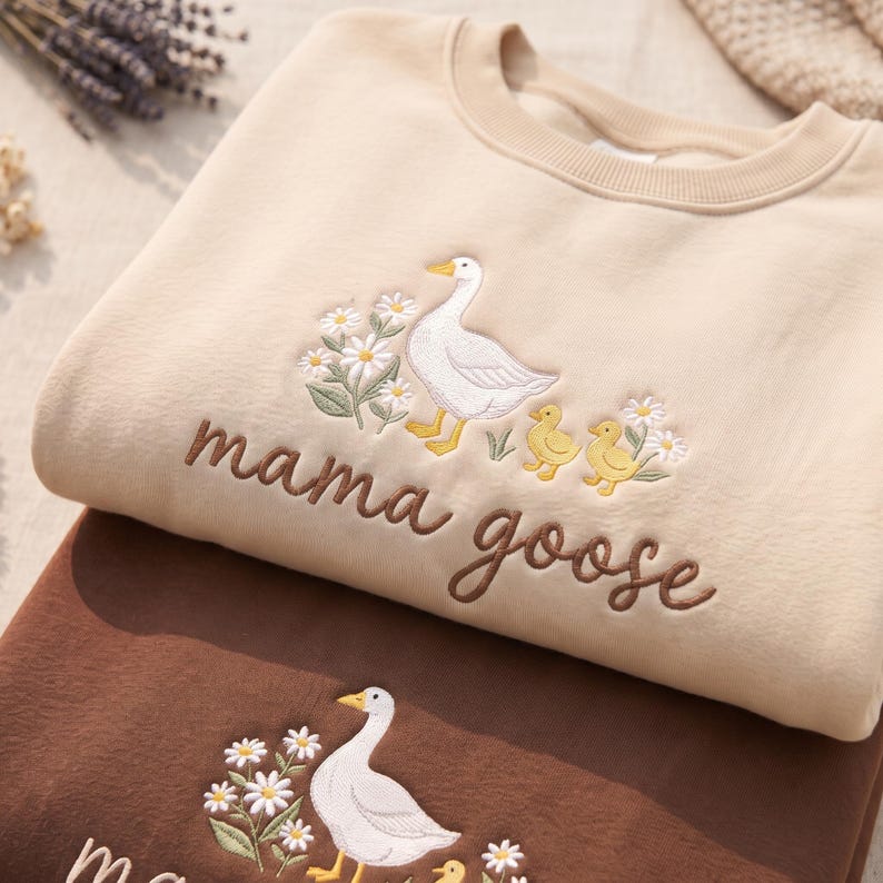 May include: A beige crewneck sweatshirt featuring an embroidered design of a white goose, two yellow goslings, and white daisies with green stems. The text "mama goose" is embroidered in brown script below the goose and goslings illustration.