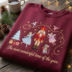 May include: A maroon crewneck sweatshirt featuring embroidered holiday designs including a nutcracker, a ballerina, a fairy, a mouse, a Christmas tree, and gifts. The text "The most wonderful time of the year" is embroidered in gold script below the figures.