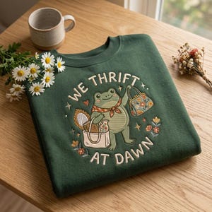 Puede incluir: Una sudadera de cuello redondo verde oscuro con un bordado de una rana con pañuelo, que sostiene un bolso floral y una bolsa de tela con un espejo y un jarrón. El texto "WE THRIFT AT DAWN" aparece arqueado alrededor del diseño.