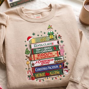 Könnte beinhalten: Ein beigefarbenes Sweatshirt mit Rundhalsausschnitt und einer Stickerei, die einen Stapel Bücher mit Weihnachtsfilmtiteln wie Elf, The Santa Clause, How The Grinch Stole Christmas, Rudolph, A Christmas Story, Christmas Vacation und Home Alone zeigt.