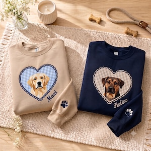 May include: A beige sweatshirt featuring a golden retriever in a blue gingham heart and a navy blue sweatshirt featuring a brown dog in a beige gingham heart. Both sweatshirts have embroidered names, Maxi and Helios, and a paw print on the sleeve.