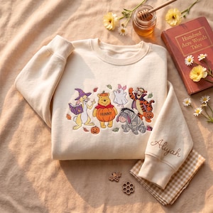 May include: A cream-colored crewneck sweatshirt featuring an embroidered graphic of Winnie the Pooh characters dressed in Halloween costumes, including a pumpkin-clad Pooh, with the name Aliyah embroidered in cursive on the sleeve cuff.