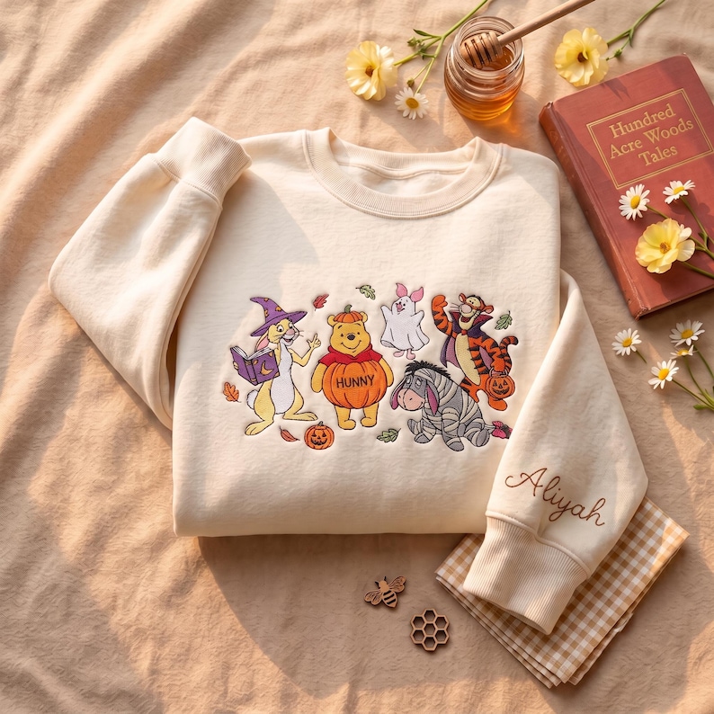 May include: A cream-colored crewneck sweatshirt featuring an embroidered graphic of Winnie the Pooh characters dressed in Halloween costumes, including a pumpkin-clad Pooh, with the name Aliyah embroidered in cursive on the sleeve cuff.