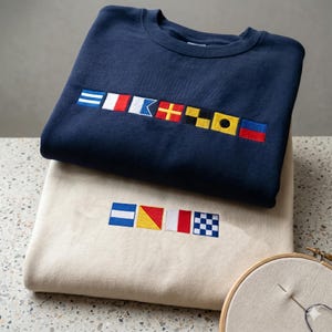 May include: A navy blue sweatshirt featuring a row of seven embroidered nautical signal flags across the chest, stacked atop a cream-colored sweatshirt with a row of four embroidered nautical signal flags centered on the front chest area.
